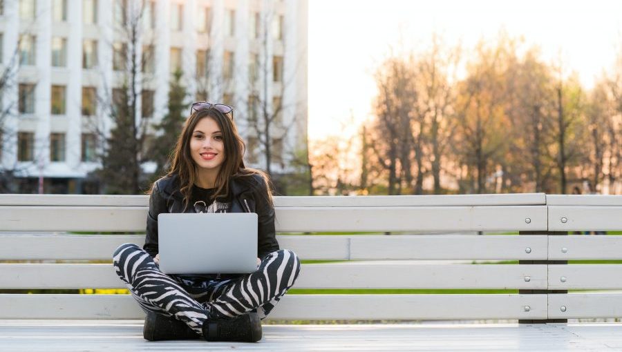 Student sitting on bench listening to music and using laptop against university campus